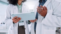 Two clinicians in lab coats review data on a tablet together while discussing a patient's care