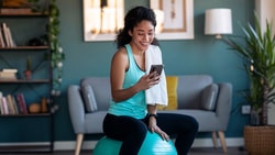 A woman in workout clothes sits on an exercise ball, smiling at her phone, in a cozy living room setting.