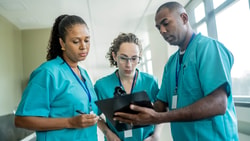 Three healthcare workers in scrubs review a clipboard, discussing training and medbridge patient care software in a hallway.