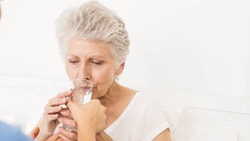 Clinician assists an older adult woman with dysphagia in drinking water during a bedside swallow assessment.