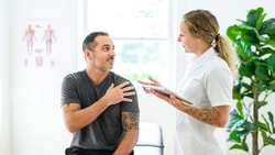 A female clinician discusses chronic shoulder pain with a male patient, applying a functional medicine framework.