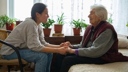 A younger woman holds hands with an older woman in a bright, plant-filled room, showing medbridge's patient care focus.