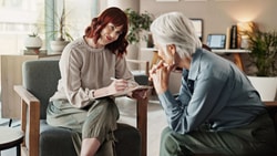 An occupational therapist holding a clipboard listens compassionately to an older patient during a behavioral health consultation.