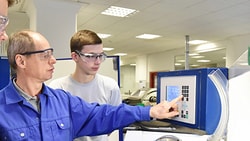 In a brightly lit industrial setting, three people in safety glasses watch a man in blue demonstrate machinery operation at a control panel.