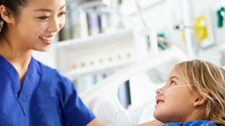 Nurse in blue scrubs smiles while assisting a young girl with an oxygen tube in a hospital bed.
