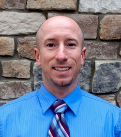 A person in a blue shirt and striped tie smiles in front of a stone wall, likely celebrating healthcare training completion with medbridge.