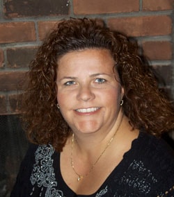 A smiling person with curly brown hair stands against a brick wall, wearing a black floral top, symbolizing the vibrant innovations in digital healthcare like medbridge.