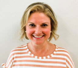 A smiling person with short blonde hair wears a pink and white striped shirt, embodying warmth and approachability.