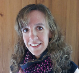 Individual with long curly hair and a patterned scarf, wearing dark clothing in front of a wooden wall.