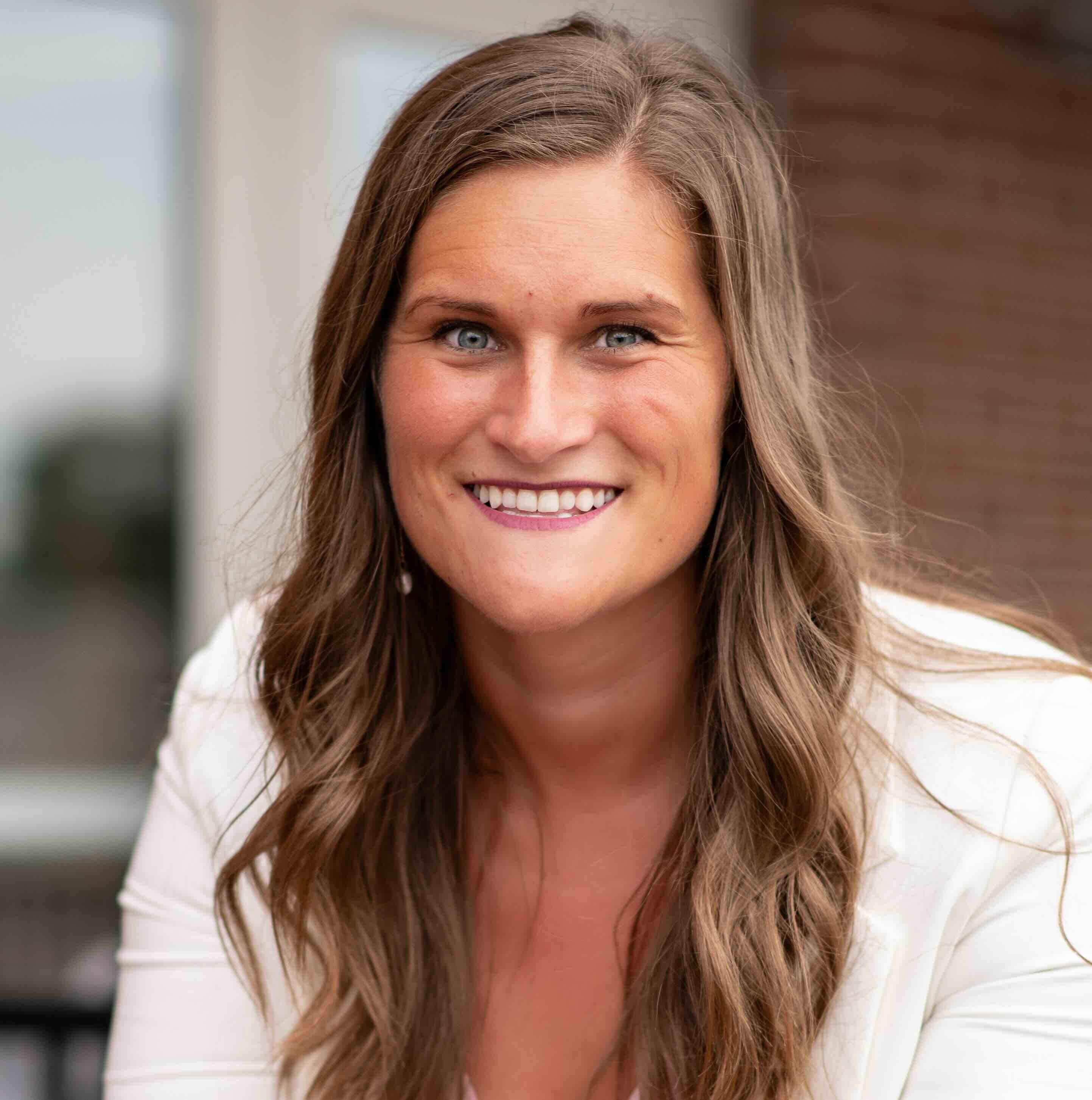A smiling woman with long wavy brown hair in a white blazer stands outdoors, embodying medbridge’s approachable healthcare.