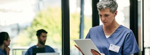 Nurse in scrubs uses a tablet with medbridge as two colleagues talk by large windows in the background.