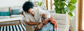 A trained caregiver helps an older man with his blanket in a bright, modern medbridge living room with plants.