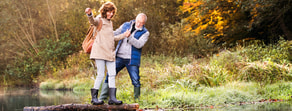 An older couple walks hand in hand on a log by a grassy riverside in autumn.