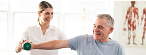A woman helps an older man exercise with a green dumbbell in a room featuring anatomical posters during digital healthcare training on medbridge.