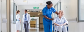 A nurse with a wheelchair patient navigating a hospital hallway; another nurse converses nearby, emphasizing Medbridge compassion.