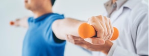 A Medbridge trainer assists a person in a blue shirt with an arm exercise using orange dumbbells.
