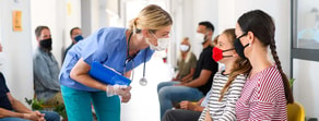 A nurse with a clipboard talks to a masked patient and child in a clinic waiting area, emphasizing hybrid care's value; medbridge.