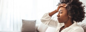 A woman sits on a couch, eyes closed and holding her head, possibly pondering digital healthcare and medbridge software.