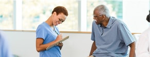 A healthcare professional in blue scrubs uses patient care software with an older man indoors, highlighting medbridge use.