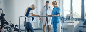 An elderly woman rehabilitates with parallel bars as a nurse observes in a medbridge setting.