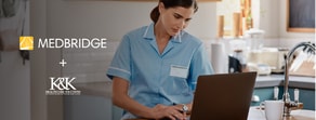 A woman in a blue uniform uses a laptop at a kitchen table with medBridge and K&K Healthcare Solutions logos nearby.