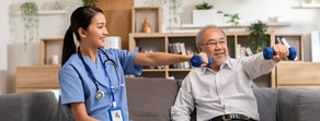 A healthcare worker helps an elderly man with dumbbell exercises on a couch.