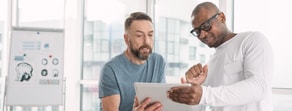 Two men in an office discuss digital healthcare software and Medbridge with a tablet, a flip chart showing brain images behind them.