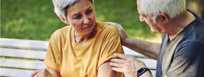 An elderly man in a gray shirt comforts a pensive woman in yellow on a park bench, reflecting medbridge’s uplifting spirit.