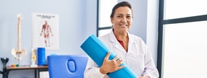 A woman in a lab coat holds a yoga mat in a bright room with Medbridge integration.