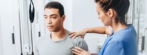 A physical therapist in blue scrubs uses medbridge tools to evaluate a man's shoulder in a clinic.