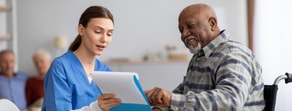 A healthcare worker in blue scrubs uses Medbridge software to discuss a document with an older man in a wheelchair.