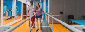 A woman with a prosthetic leg walks between parallel bars in a Medbridge hybrid care setting, assisted by a supportive man.