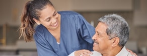 A caregiver in blue scrubs smiles at an elderly man, showcasing her medbridge healthcare training.