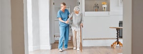 A caregiver trained through medbridge helps an elderly woman with a cane in a modern living room.