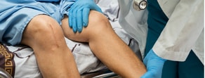A doctor in a white coat and blue gloves examines a patient's knee, showcasing medbridge's digital healthcare tools.