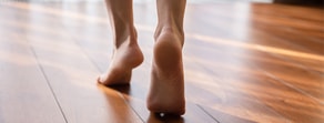 Barefoot person walking on polished wooden floor, highlighting feet and lower legs.