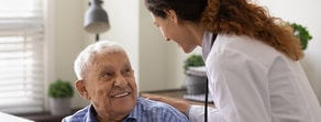 A healthcare professional with a white coat and long curly hair smiles warmly at an elderly man, showcasing Medbridge's digital healthcare benefits.