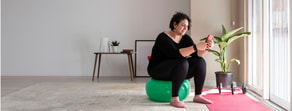 A person on an exercise ball checks their phone in a cozy room with fitness gear, reflecting digital health trends.