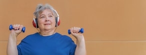 An older adult with gray hair exercises with blue dumbbells, wearing headphones, against an orange wall, embodying Medbridge's healthcare education benefits.