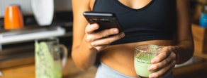 A person in a sports bra uses their smartphone, possibly accessing medbridge, while holding a green smoothie in the kitchen.