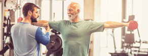 An elderly man works out with dumbbells, guided by a younger trainer in the gym, embodying medbridge's commitment to lifelong strength and vitality.