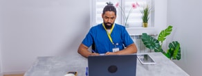 A man in blue scrubs works at a desk with a laptop, tablet, book, and paper; potted plants are behind him.
