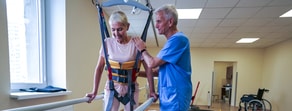 A physical therapist helps a woman in a harness use parallel bars, with healthcare education subtly provided, while a wheelchair is in the background.