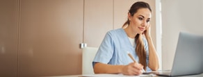 A healthcare worker in blue scrubs uses medbridge software on a laptop while writing at a desk.