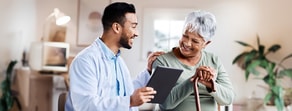 A healthcare worker uses a tablet with medbridge software while interacting with a smiling elderly woman.