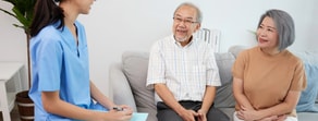 A healthcare professional speaks with an older couple on a couch in a bright room.