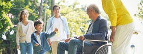 A child runs toward a person in a wheelchair, joined by adults, in a sunny park.