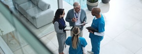 Four professionals in medical and business attire discuss Medbridge patient care software with folders and tablets in a modern setting.
