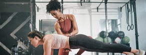 A fitness trainer helps a woman with her plank exercise in a gym, using medbridge principles.