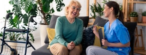 An elderly woman talks with a smiling healthcare worker about patient care software, sitting on a sofa near a walker and plants.